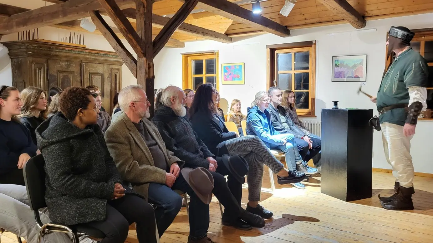 Eine Gruppe von Menschen sitzt in einem hellen Raum mit Holzbalken und lauscht aufmerksam einem Mann in mittelalterlicher Kleidung, der vorne steht und spricht.