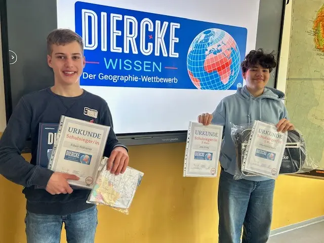 Zwei Schüler der Regelschule Schmalkalden zeigen ihre Urkunden und Preise beim Geographiewettbewerb Diercke Wissen 2026 vor einem Bildschirm mit Wettbewerbslogo.