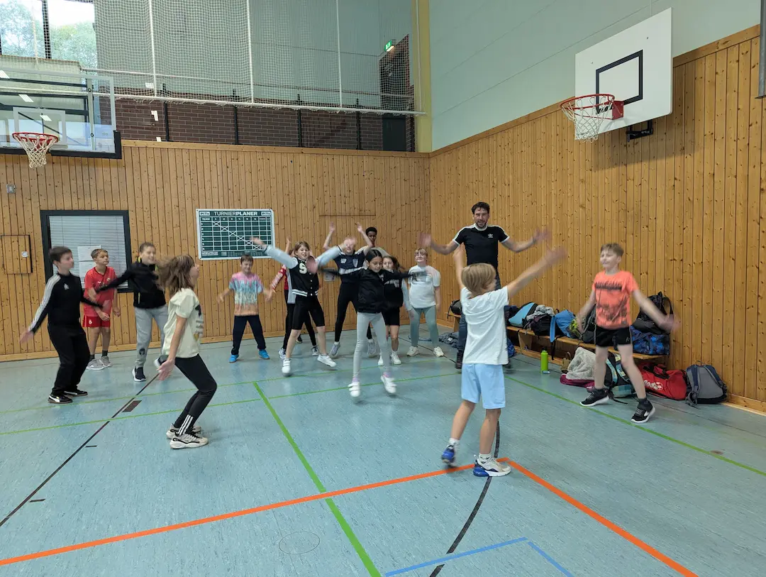 Kinder und Jugendliche wärmen sich in einer Turnhalle auf. Sie machen Hampelmänner, während ein Trainer vorne die Übungen anleitet.